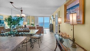 A spacious living area with a dining table, chairs, and a large plant. There is a sofa and a TV in the background.