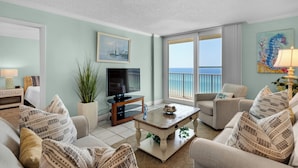 A spacious living room with a television, comfortable seating, a coffee table, and a large window overlooking the ocean.