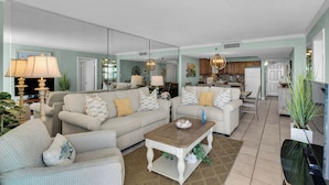 A spacious living room with a large sofa set, a coffee table, and a dining area with a kitchen in the background.