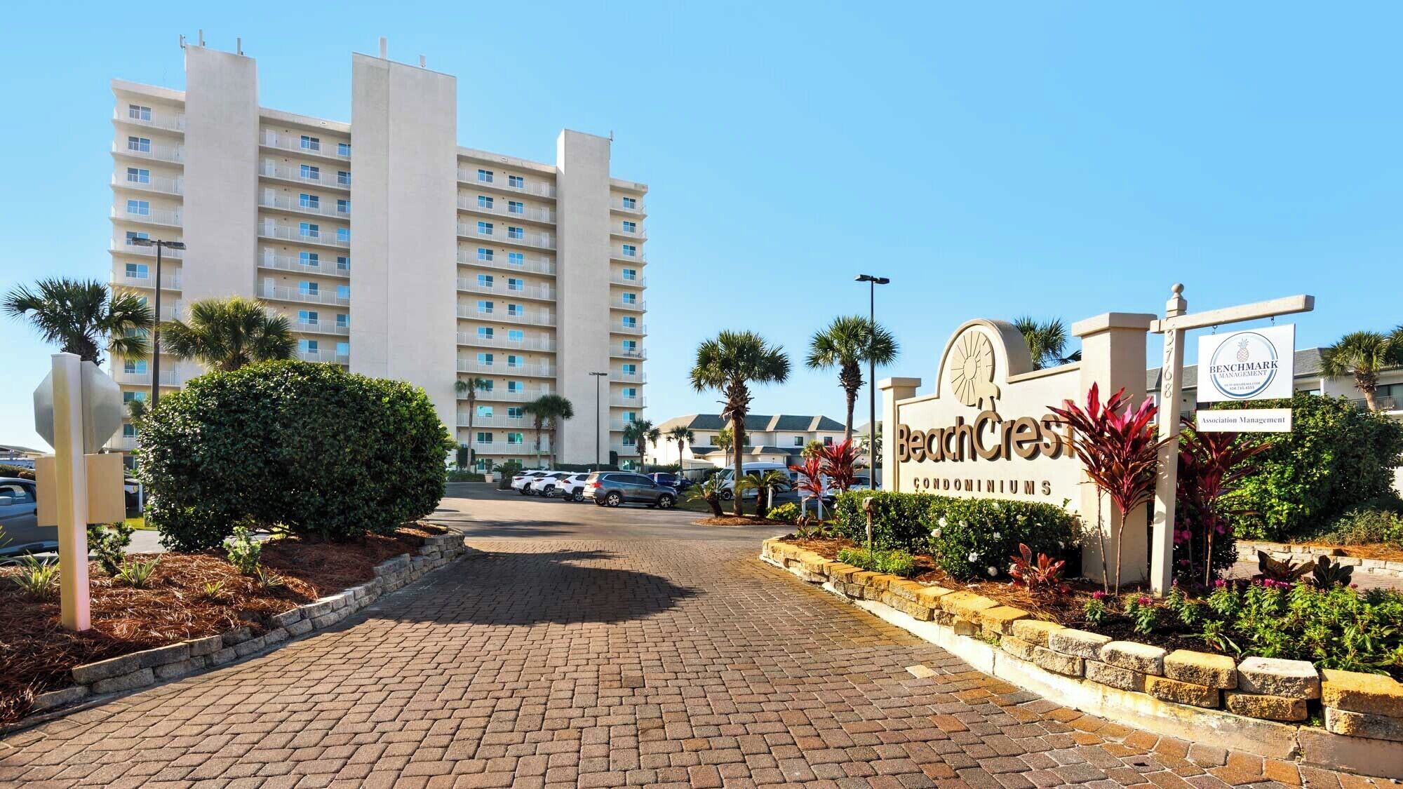 Un complexe de condominiums situé en bord de mer, doté d’une allée pavée, de palmiers et d’un panneau indiquant « BeachCrest Condominiums ».