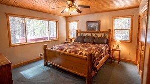 A wooden-framed bedroom with a bed, two bedside tables, a ceiling fan, and large windows.