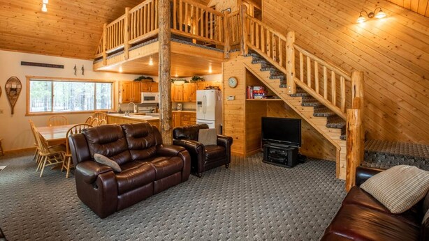 A spacious living area with a leather sofa, a dining table with chairs, a kitchen with wooden cabinets, and a wooden staircase.