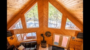 Ein gemütliches Blockhaus-Interieur mit Holzbalkendecke, Kamin, einer Couch und großen Fenstern mit Blick auf den umliegenden Wald.