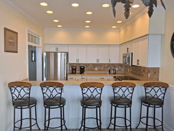 A modern kitchen with white cabinets, a stainless steel refrigerator, a microwave, and a coffee maker. There are five bar stools with metal frames and cushioned seats.
