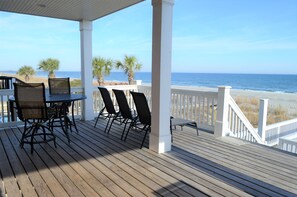 A wooden deck with a table and chairs, overlooking a beach with palm trees and the ocean.
