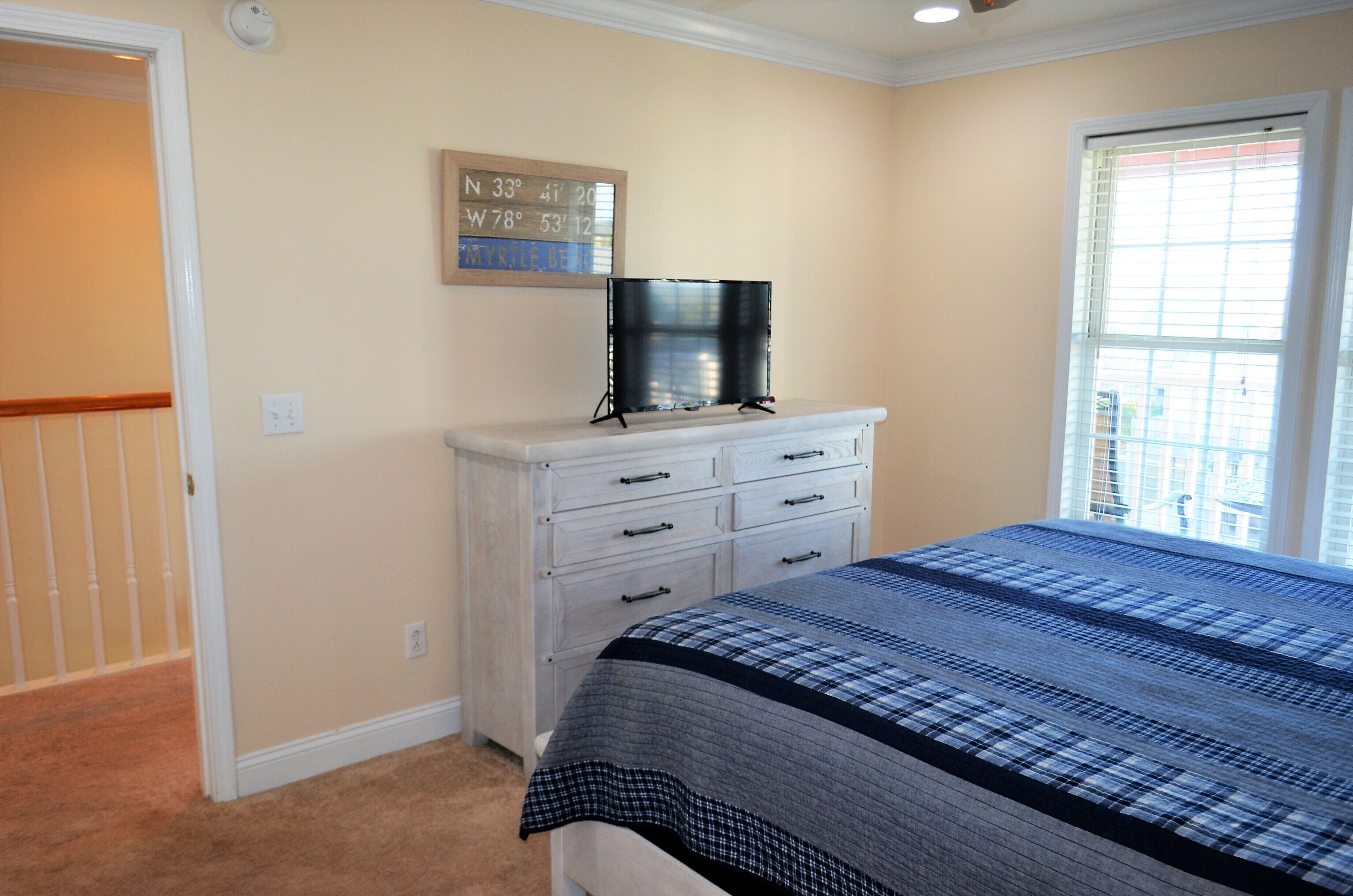 A bedroom with a bed, a dresser, a flat-screen TV on the dresser, and a window with blinds.