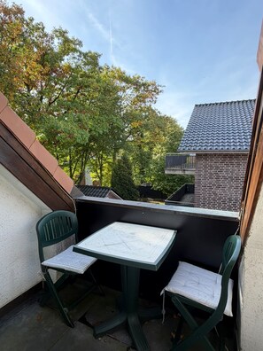 Ein Balkon im Freien mit Tisch und zwei Stühlen, umgeben von Bäumen und einem Gebäude mit einem Ziegeldach.