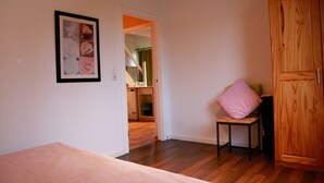 Huoneessa on sänky, vaaleanpunaisella tyynyllä varustettu tuoli, puinen vaatekaappi ja seinällä oleva kehystetty kuva.