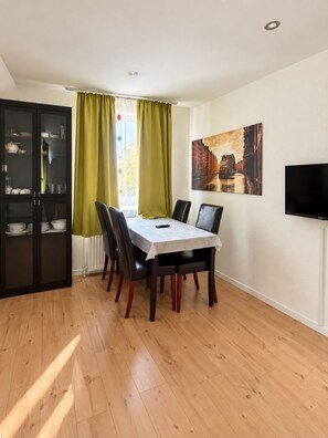 Uma sala de jantar com mesa e cadeiras, um armário com louças, uma janela com cortinas e um quadro na parede.