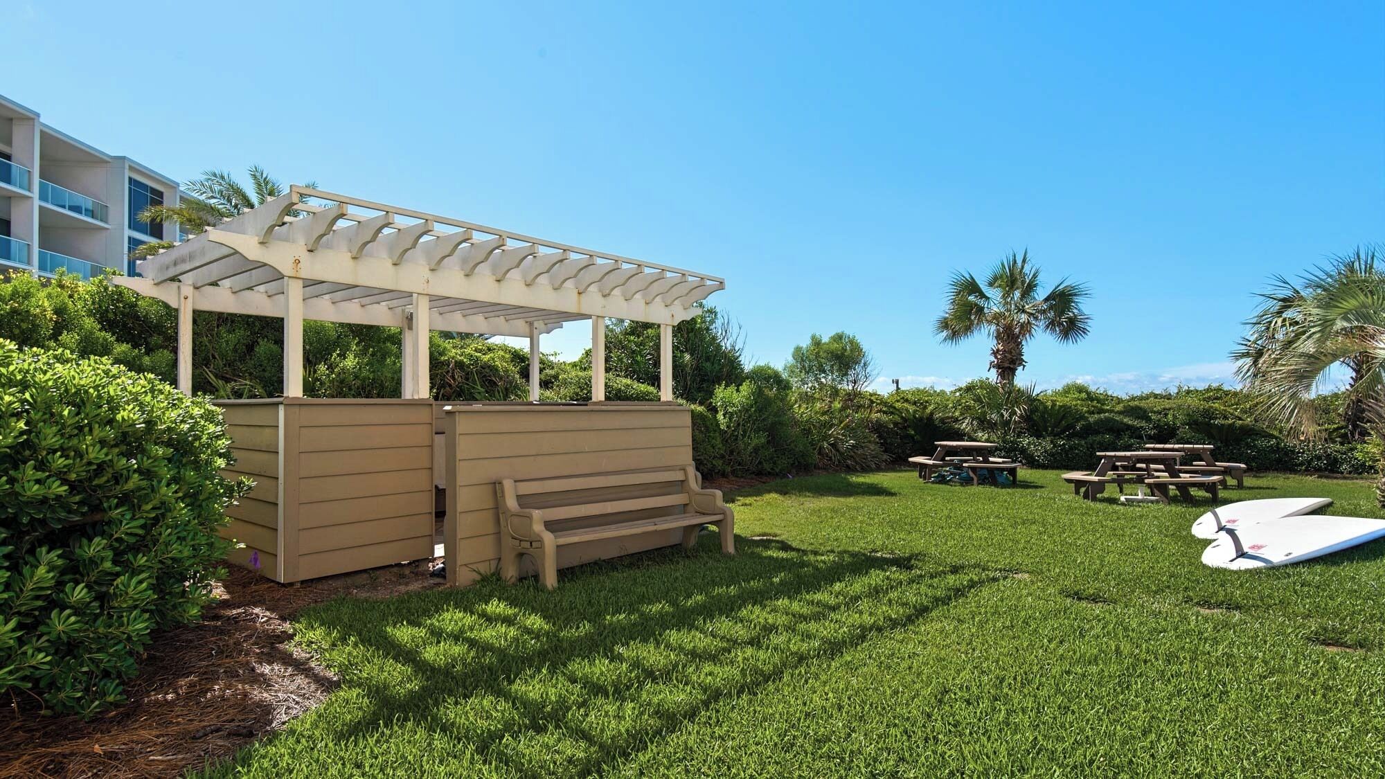 A well-maintained outdoor area with a wooden pergola, a bench, picnic tables, and a surfboard.