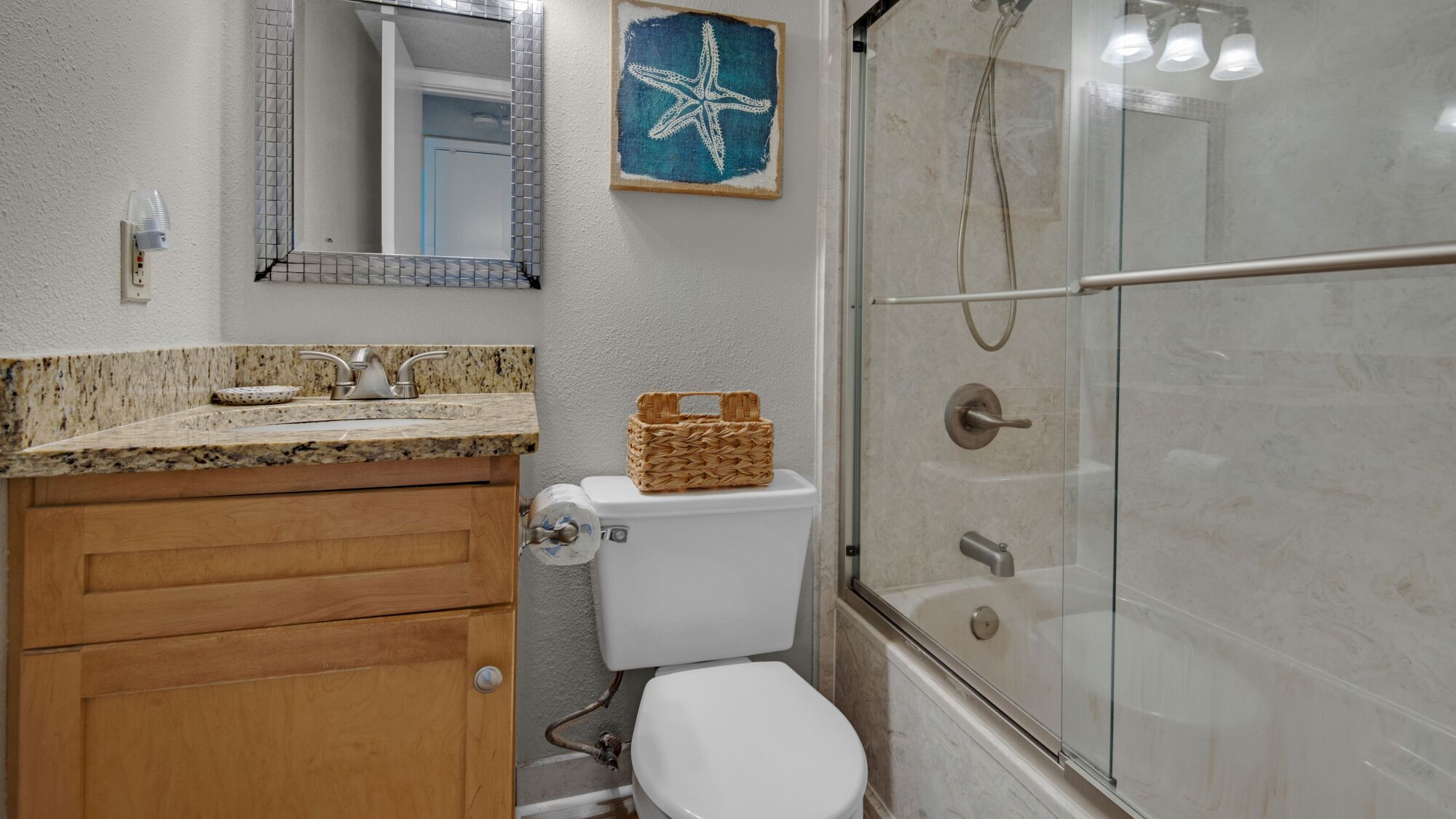 Une salle de bain comprenant des toilettes, un meuble en bois, un miroir, une œuvre d’art représentant une étoile de mer et une douche fermée par des parois de verre.