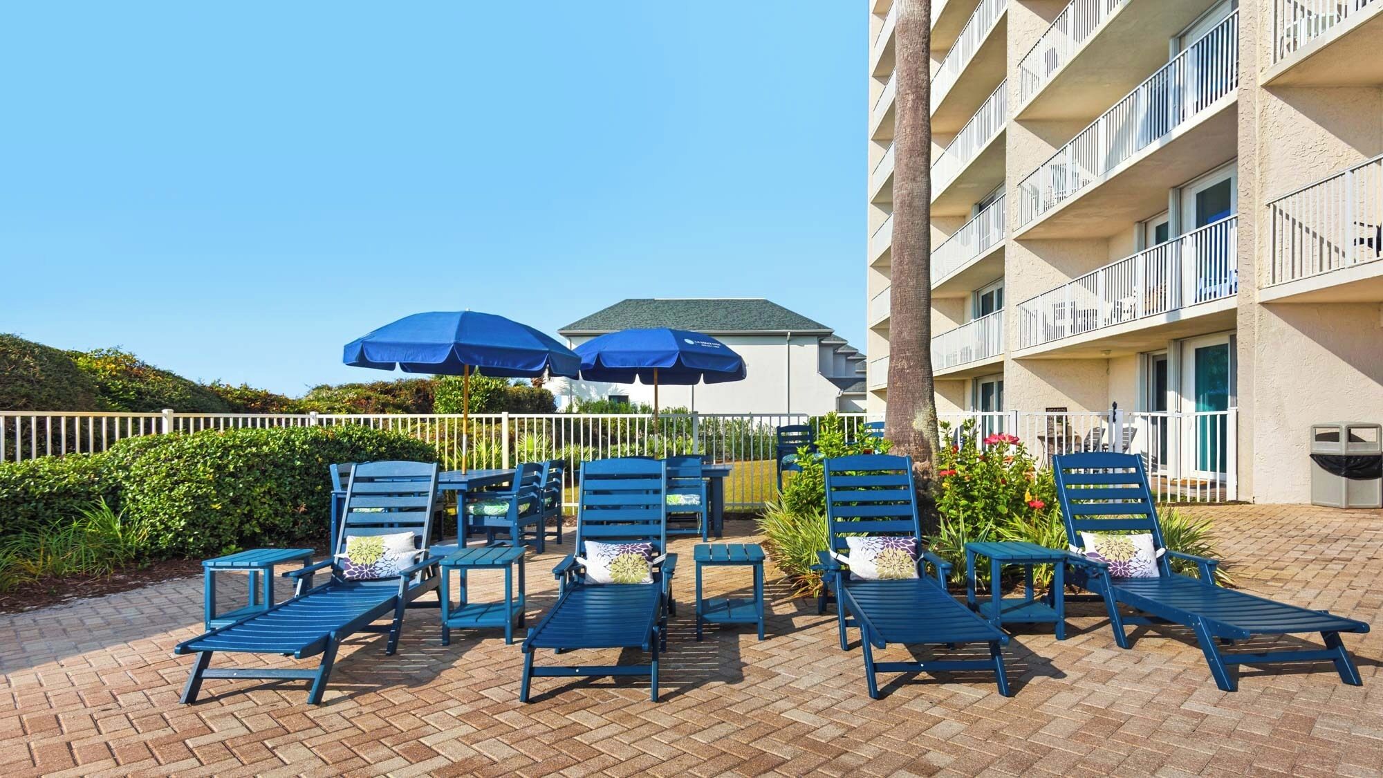 A rooftop lounge area with blue lounge chairs and umbrellas, a white fence, and a building with balconies in the background.