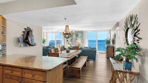 A spacious living area with a large window overlooking the ocean, a wooden dining table, and a cozy seating area. - 12th Floor View w/ Private Beach, Beach Chairs + Attraction Tickets! (Santa Rosa Beach)