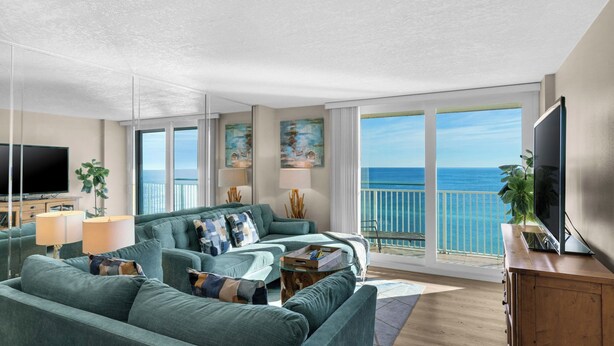 A modern living room with a large sofa, a television, and a balcony with a view of the sea. - 12th Floor View w/ Private Beach, Beach Chairs + Attraction Tickets! (Santa Rosa Beach)