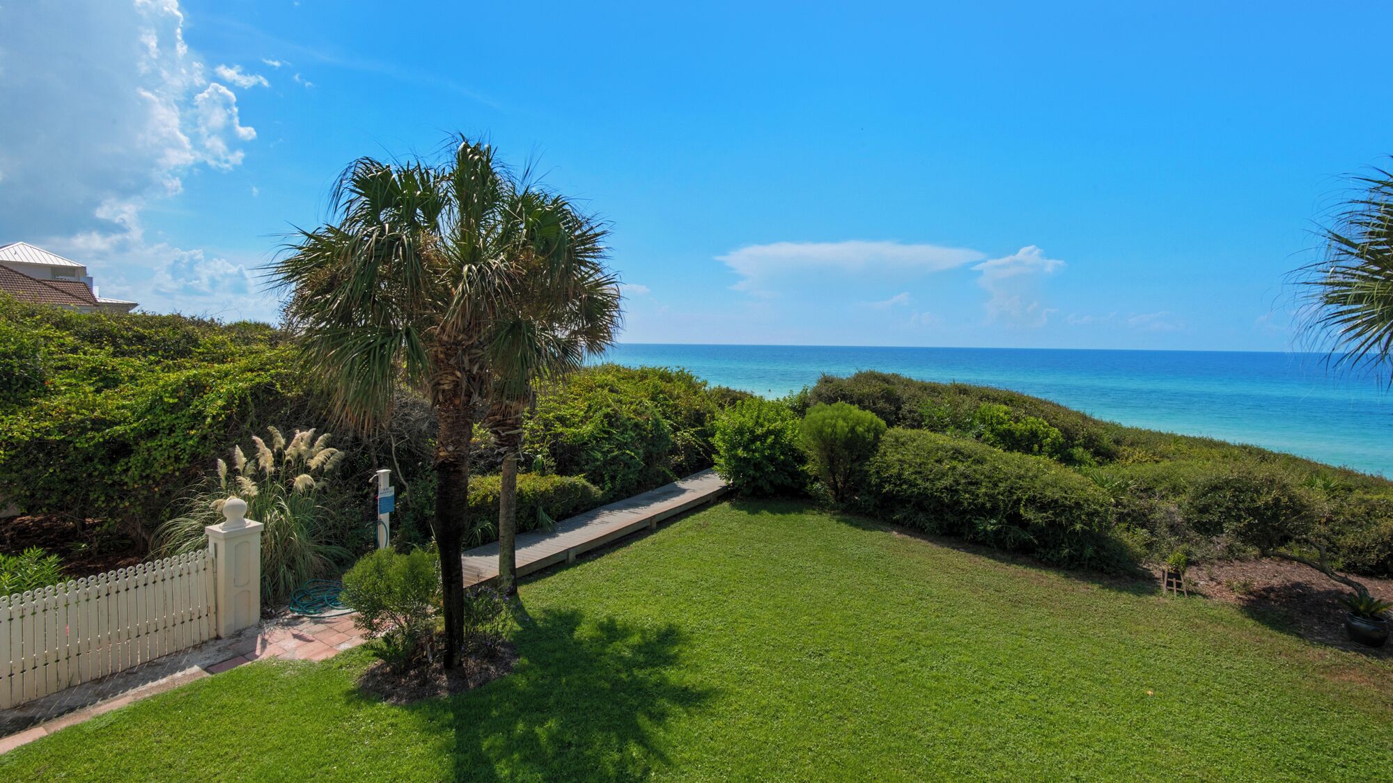 A well-maintained lawn with a pathway leading to a beach, surrounded by lush greenery and a clear view of the ocean.