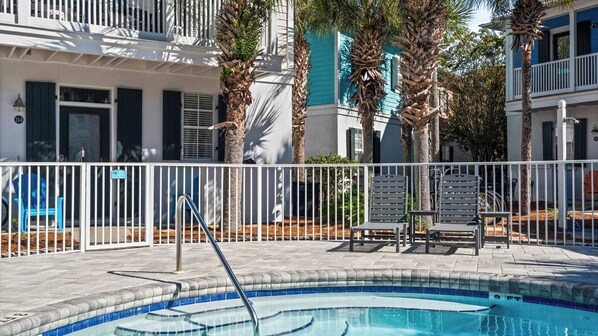 A pool area with a white fence, lounge chairs, and a building with blue shutters and palm trees.