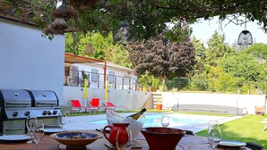 An outdoor dining area with a pool, a grill, and a view of the surrounding garden.