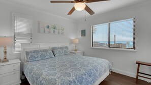 Um quarto com cama, mesa de cabeceira, abajur, ventilador de teto, janela com persianas e vista para o oceano.