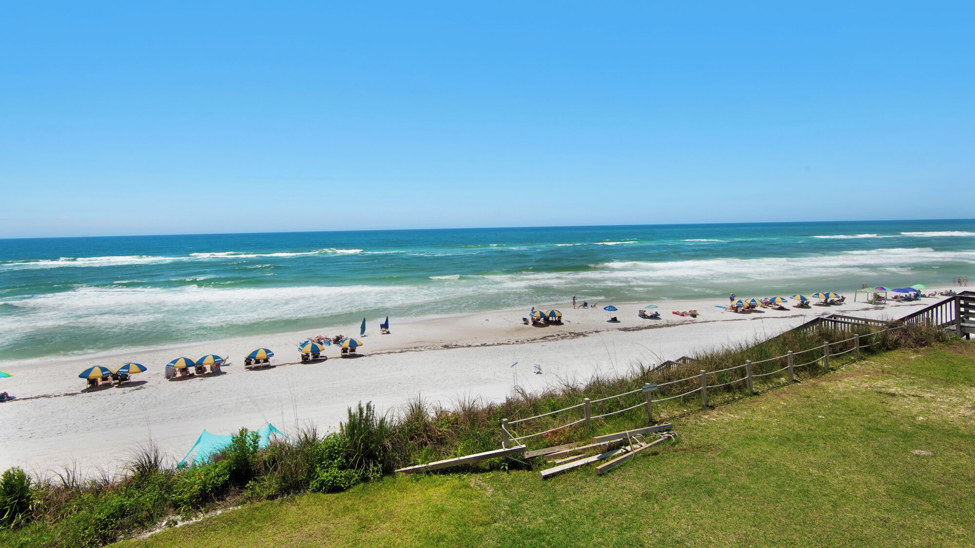 A beach with umbrellas and beach chairs, a wooden fence, and a grassy area.
