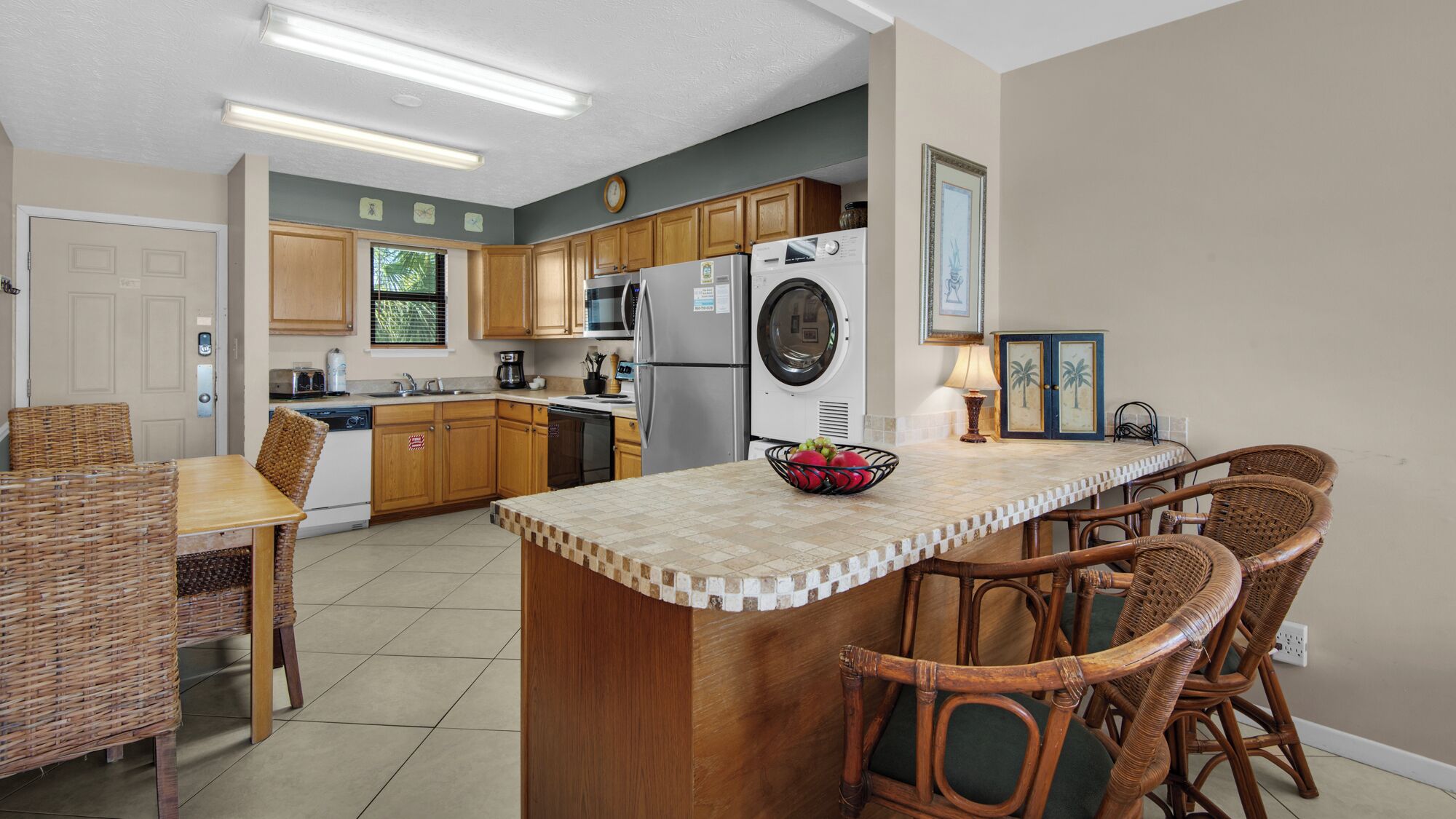 A kitchen with wooden cabinets, a built-in washing machine, a dining area with wicker chairs, and a tiled backsplash.