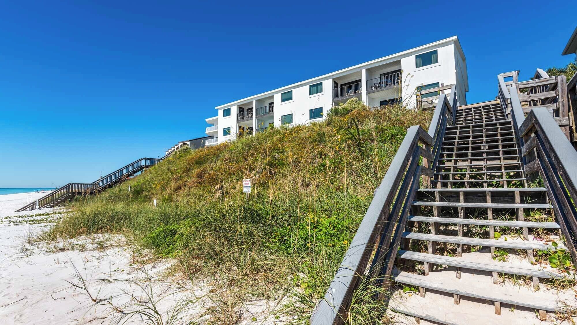 Un edificio moderno frente a la playa con una escalera que lleva a un balcón, rodeado de vegetación costera.