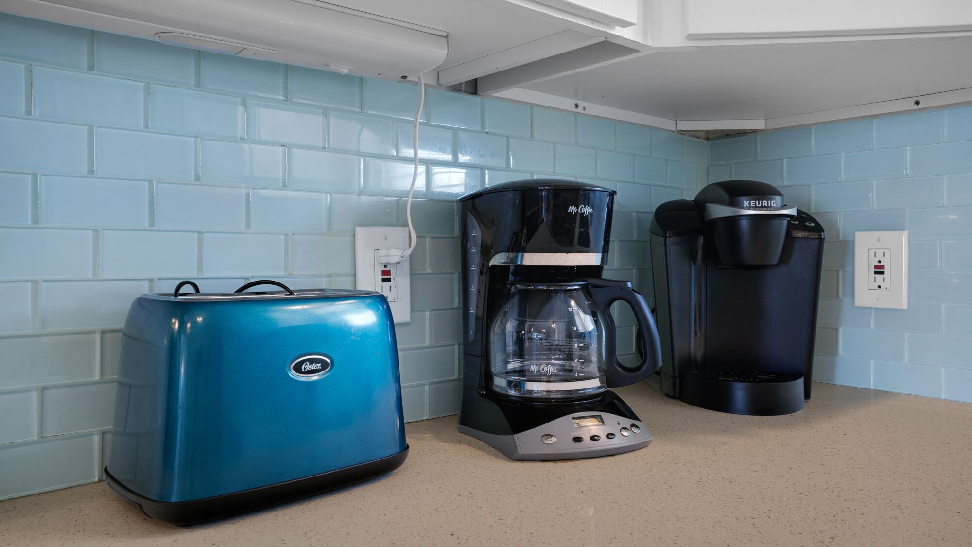 A kitchen with a blue Oster toaster, a Mr. Coffee coffee maker, and a Keurig coffee maker on a beige countertop.