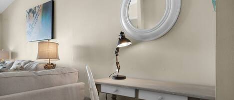 Un dormitorio con cama, escritorio con lámpara, espejo y un cuadro en la pared.