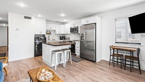 A modern kitchen with white cabinetry, stainless steel appliances, a central island with stools, and a dining area with a wooden table and chairs. - 1st Floor, Gulf Side in Seagrove,  Sleeps 6 + Free Attraction Tickets! (Santa Rosa Beach)