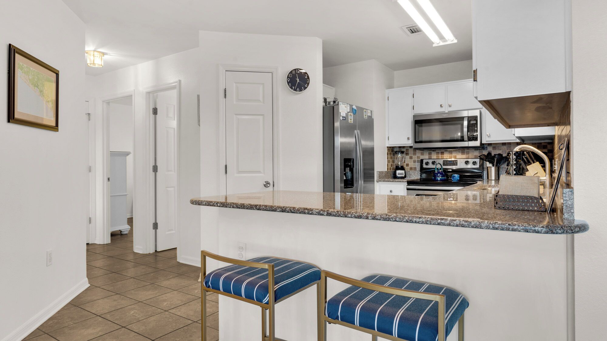 A modern kitchen with white cabinetry, a stainless steel refrigerator, a microwave, and an oven. There are two bar stools with blue cushions.