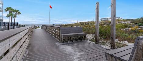 Un passeig de fusta que condueix a una platja amb una bandera vermella, una banca i una construcció de fusta al fons.