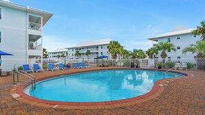 A swimming pool surrounded by a brick patio, with lounge chairs and umbrellas, and a multi-story building with balconies in the background.
