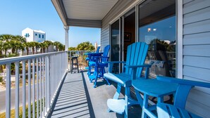 A covered balcony with blue Adirondack chairs and a table, overlooking a street with palm trees and a white building in the background.