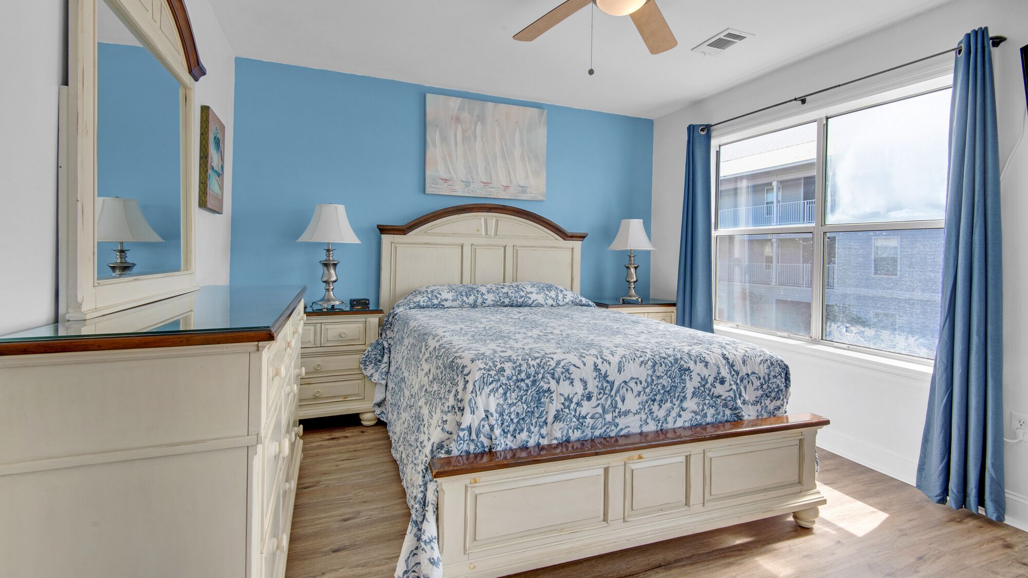 Une chambre à coucher comprenant un lit, des tables de chevet, une commode, un ventilateur de plafond et une grande fenêtre avec des rideaux.