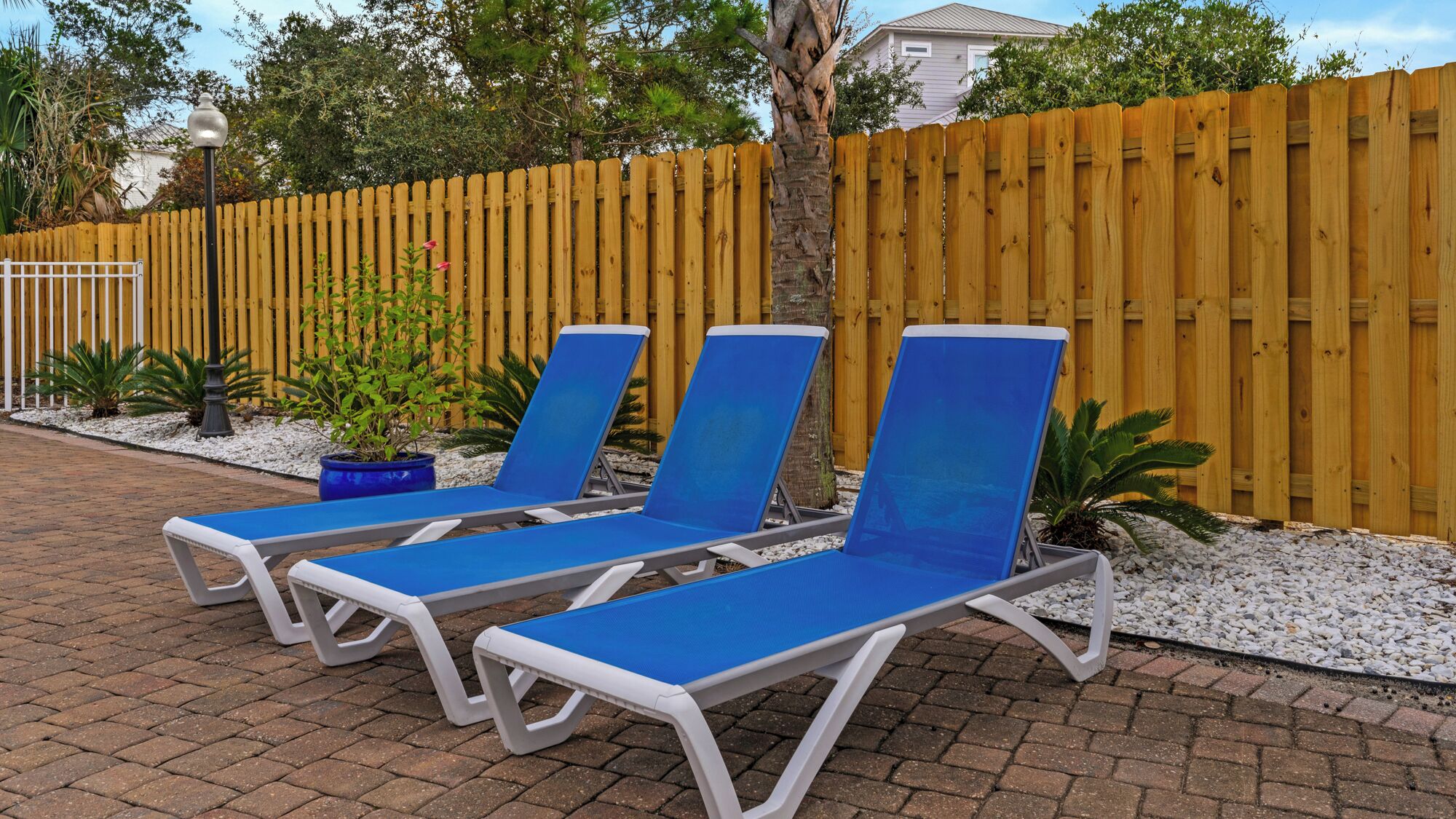A row of blue lounge chairs with white frames, positioned on a brick pathway with a wooden fence and potted plants in the background.
