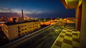 Vista noturna de uma varanda com piso xadrez, com vista para um horizonte urbano com edifícios e uma montanha ao longe.