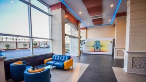 Um hall de entrada com balcão de recepção, assentos em azul e amarelo, uma grande janela com vista para carros estacionados e palmeiras, e uma parede com uma pintura de cactos.