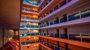 Interior de hotel com vários níveis, átrio central, guarda-corpos de vidro e padrão de piso xadrez.