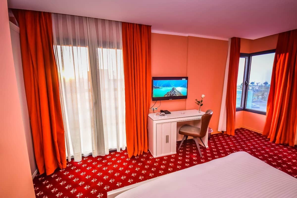 Una habitación de hotel con cama, escritorio, televisor y ventana con cortinas.