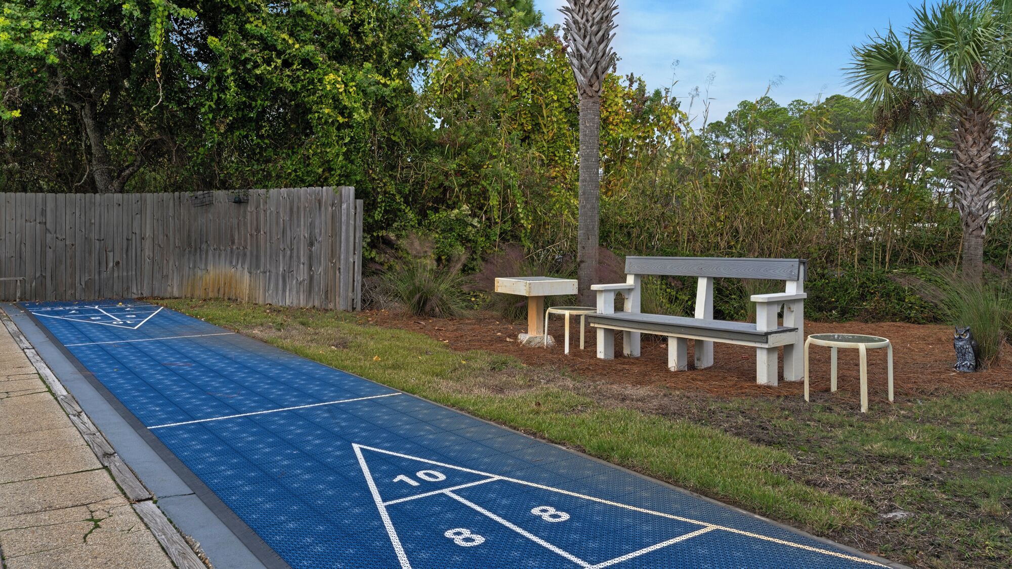 A shuffleboard court with a wooden fence, a bench, and an owl statue.