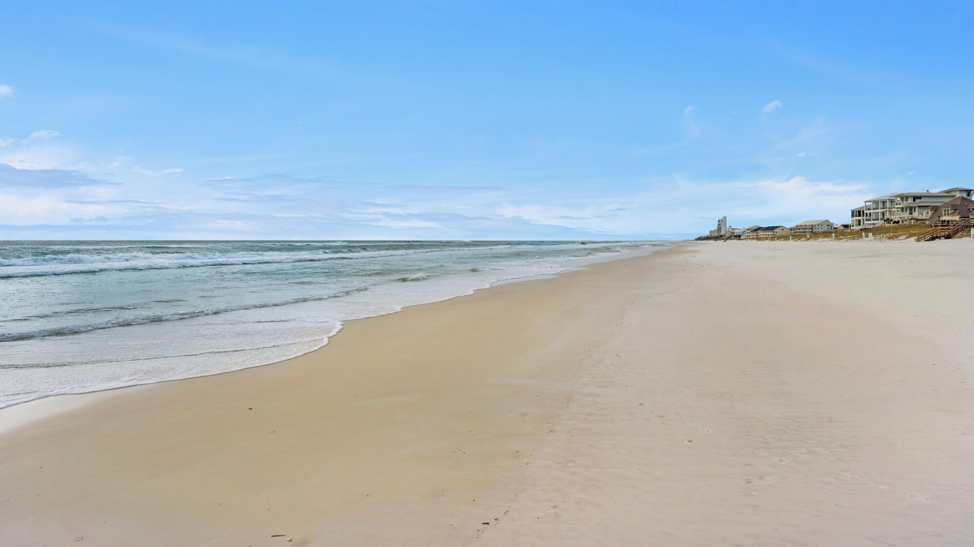 A beach with a clear sky, a sandy shore, and a building with multiple stories and balconies.