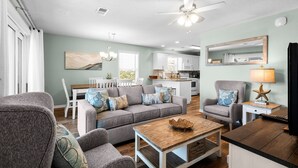Ein Wohnzimmer mit grauem Sofa, einem Holztisch und einem grauen Sessel. Im Hintergrund befindet sich eine Küche mit weißen Schränken und ein Essbereich mit weißem Tisch und Stühlen.