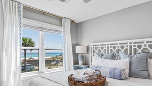 Ein Schlafzimmer mit Blick auf den Strand, ein Bett mit weißer Bettwäsche, ein Korbwäschesystem und ein Nachttisch mit einer Lampe.