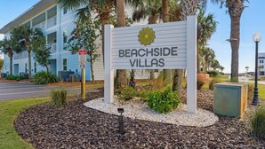 Señal de Beachside Villas con logotipo de flor, rodeada de jardines y un edificio al fondo.