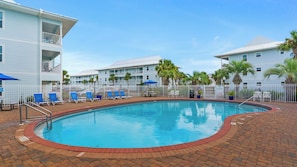A swimming pool with lounge chairs and umbrellas, surrounded by a brick walkway and white fences.