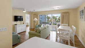 A room with a flat-screen TV, a sofa, a dining table with chairs, a small kitchenette, and a balcony with a view of palm trees and buildings. - Steps from the Gulf ~ 2 Community Pools ~ Tennis + Free Attraction Tickets! (Santa Rosa Beach)