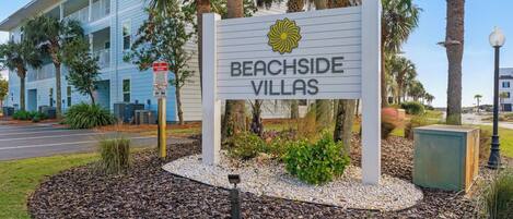 A sign for Beachside Villas with a flower logo, surrounded by landscaping and a building in the background.
