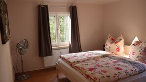 A bedroom with a bed, a standing fan, a window with curtains, and a radiator. - New apartment in the middle of vineyards (Coswig)