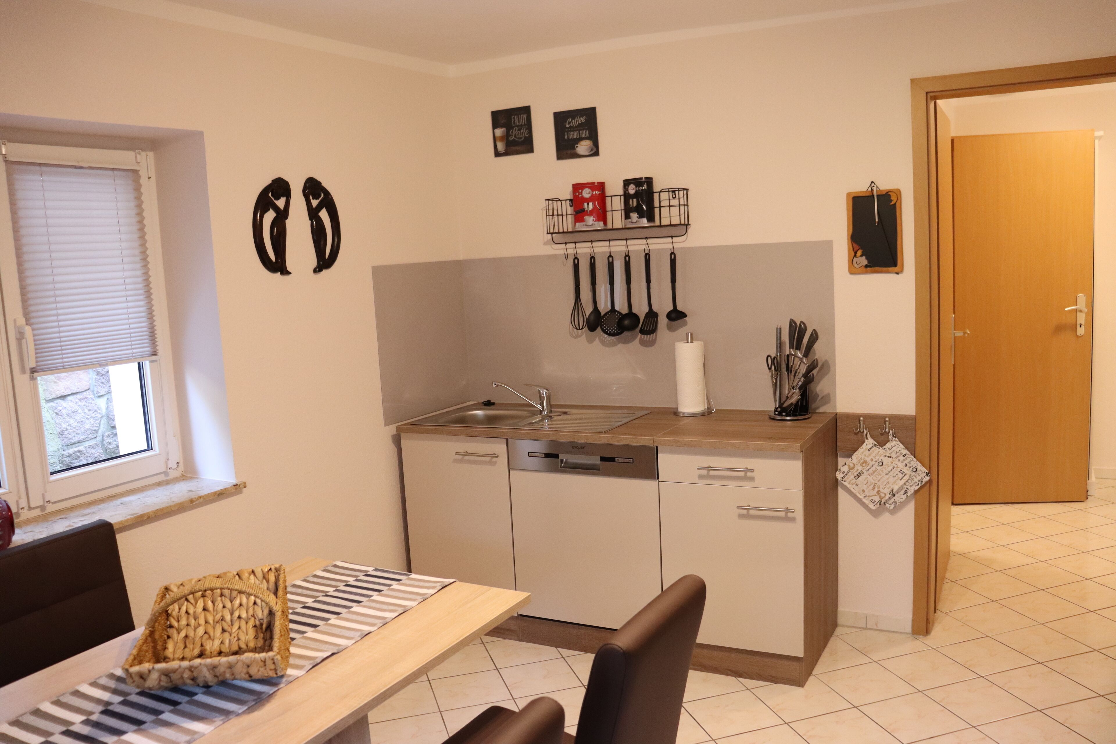 A compact kitchen with a sink, dishwasher, and a small countertop. There are two wall-mounted hooks above the sink, and a window with blinds to the left.