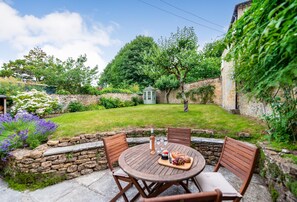 A patio with a round wooden table and four chairs, surrounded by a stone wall and lush greenery.