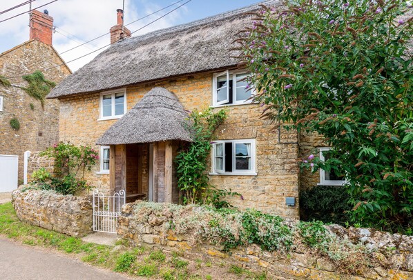 A stone house with a thatched roof, a wooden door, and a small garden with flowering plants.